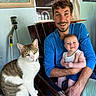 man, baby, cat, chair, wood, window, painting, indoor, smile, happy, person, animal, pet, holding, casual_clothing, green_eyes, baby_bottle, decor, furniture, portrait