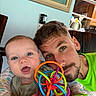 baby, man, toy, colorful_toy, blue_eyes, beard, green_shirt, highchair, indoor, smiling, closeup, face, portrait, family, parent, child, happy, wooden_chair, painting, window