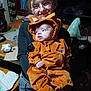 child, baby, costume, orange, glasses, smile, indoor, person, holding, table, furniture, clothing, face, cute, happy, window, plaid_pants, headwear, home, sibling