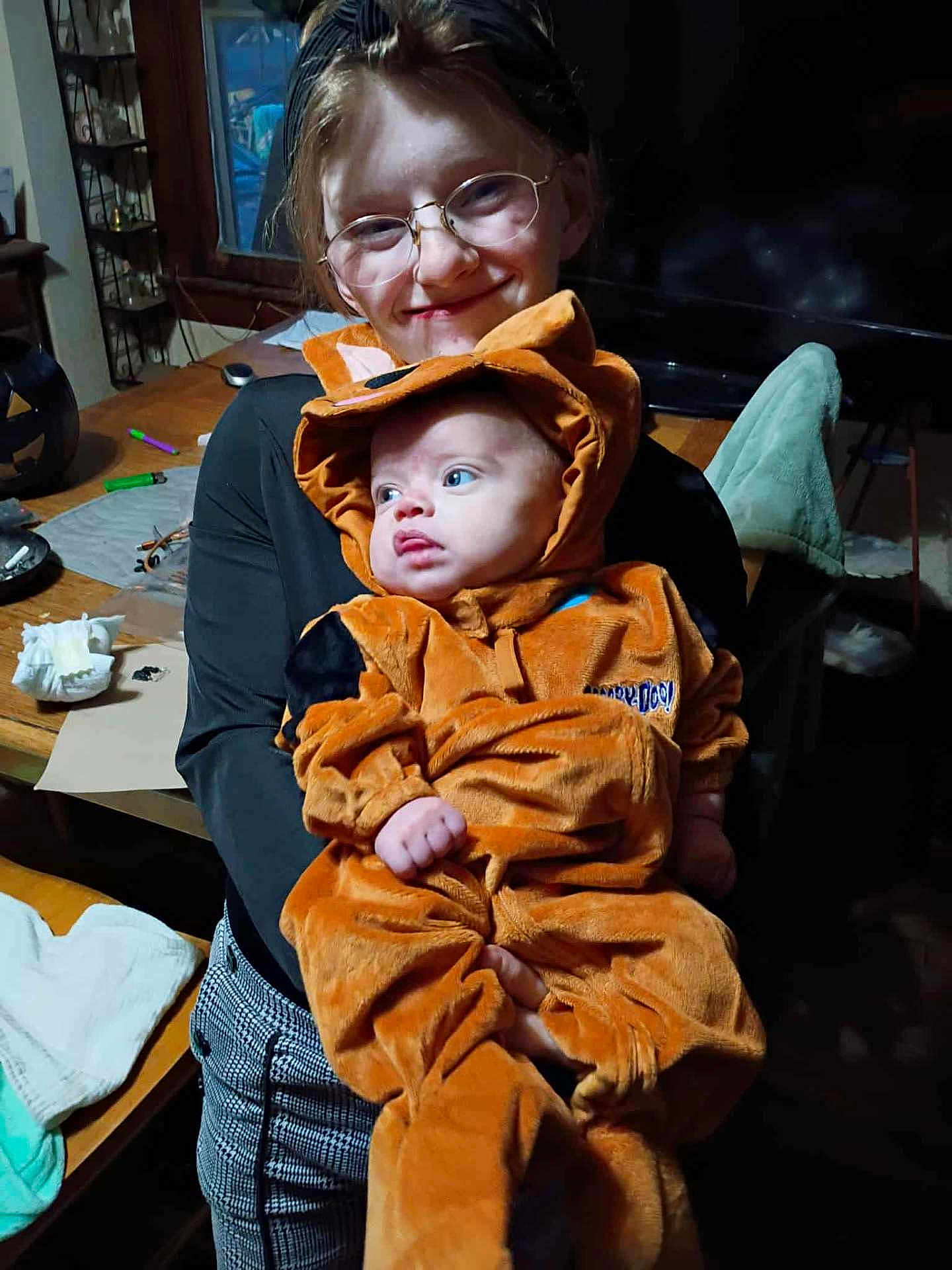 Ryker is registered to the contest to win money with this photo: child, baby, costume, orange, glasses, smile, indoor, person, holding, table, furniture, clothing, face, cute, happy, window, plaid_pants, headwear, home, sibling