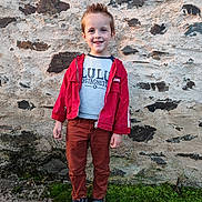 Léo a rejoint le concours — aidez-le/la à gagner de superbes lots ! boy, brown_pants, casual_clothing, child, daylight, fashion, footwear, full_body, grass, hair_style, happy, nature, outdoor, person, portrait, red_jacket, smiling, sneakers, standing, stone_wall