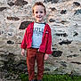 boy, brown_pants, casual_clothing, child, daylight, fashion, footwear, full_body, grass, hair_style, happy, nature, outdoor, person, portrait, red_jacket, smiling, sneakers, standing, stone_wall