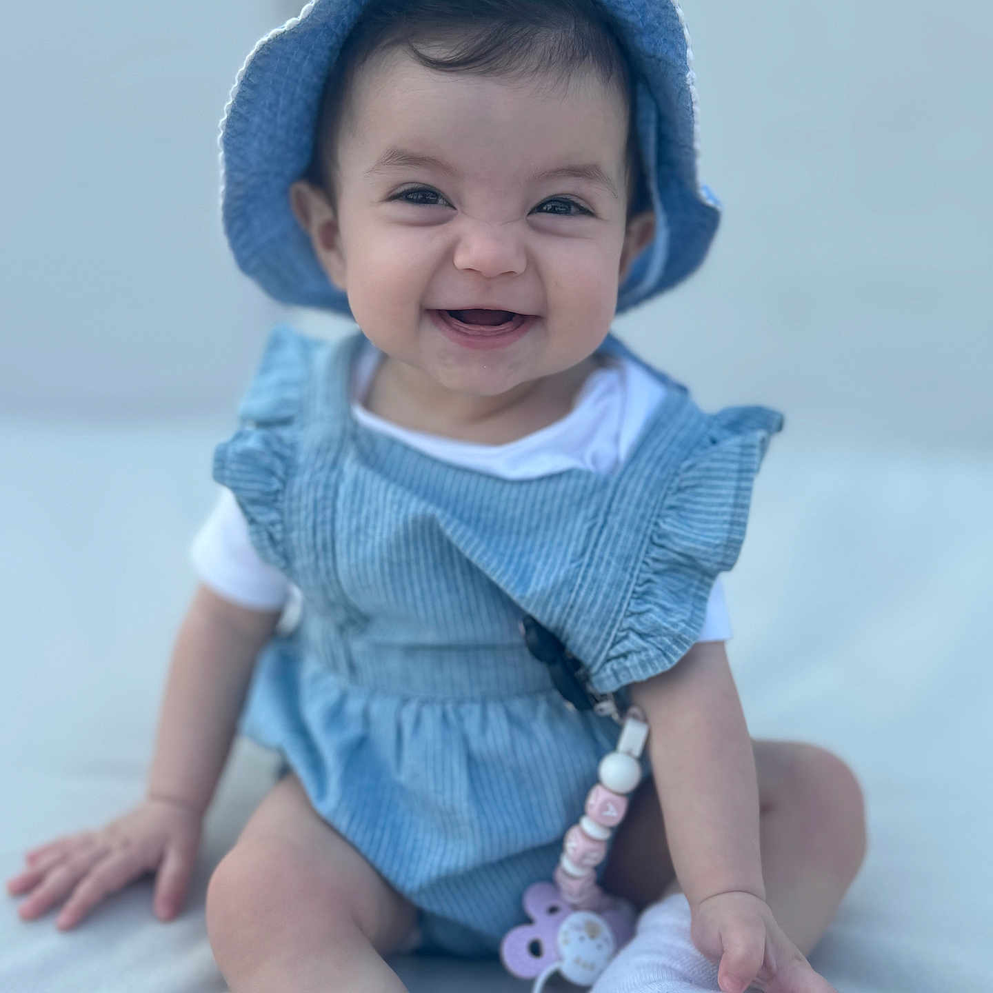 Anaya participe au concours pour gagner de l'argent avec cette photo : baby, baby_toy, blue_hat, child, cute, face, hand, happy, indoors, infant, pacifier_clip, person, playful, romper, sitting, skin, smiling, soft_background, white_socks, young_child