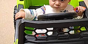 Amir joined the competition — help win amazing prizes! baby, child, toy_car, indoor, car, green, car_seat, steering_wheel, floor, carpet, person, infant, hand, clothing, sitting, expression, room, hallway, doorway, looking
