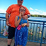 birds, blue_dress, blue_sky, casual_clothing, child, clouds, daylight, ducks, fence, girl, glasses, lake, man, nature, orange_shirt, outdoor, people, snack, summer, water