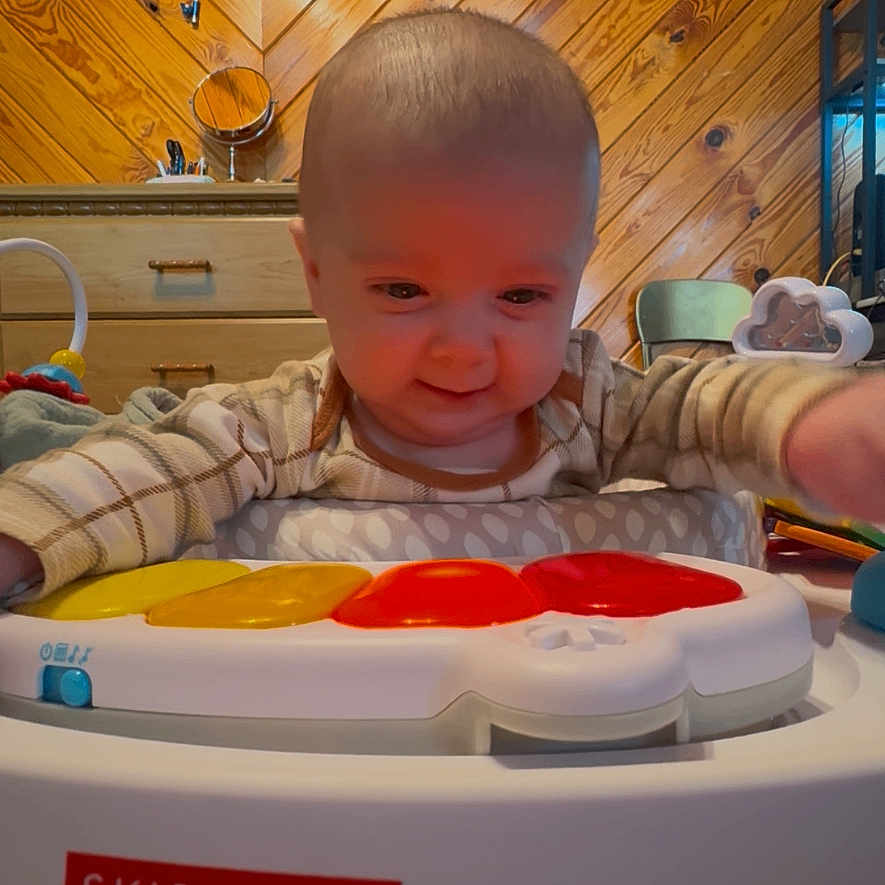 Cayde is registered to the contest to win money with this photo: baby, child, toy, skip_hop, indoor, wooden_wall, play, infant, smiling, cute, clothing, furniture, activity_center, happy, person, head, face, colorful, reaching, carpet