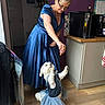 woman, dog, dress, blue_dress, tutu, indoor, kitchen, wooden_floor, playful, pet, small_dog, white_dog, standing_dog, interaction, joyful, glasses, short_hair, cabinet, countertop, appliance