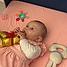 baby, child, pink_dress, bed, toy, chocolate_bunny, gold_wrapping, red_bow, teething_ring, plush_toy, soft_toy, lying_down, indoor, cute, infant, playing, pastel_colors, young_child, hand, face