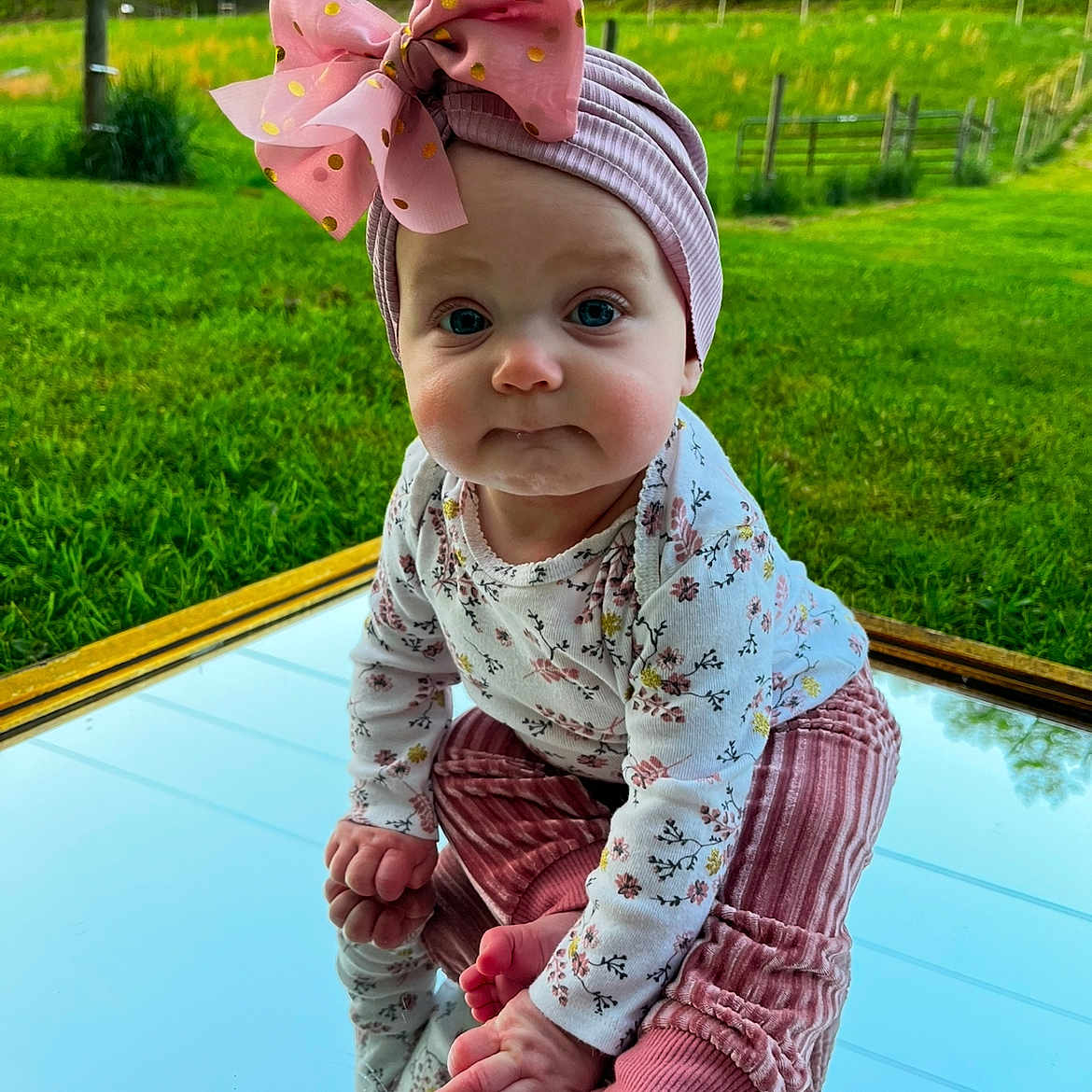 Kind is registered to the contest to win money with this photo: baby, child, clothing, cute, daylight, expression, grass, greenery, headwear, infant, mirror, nature, outdoor, pink_bow, portrait, reflection, scenic, sitting, toes, young_child