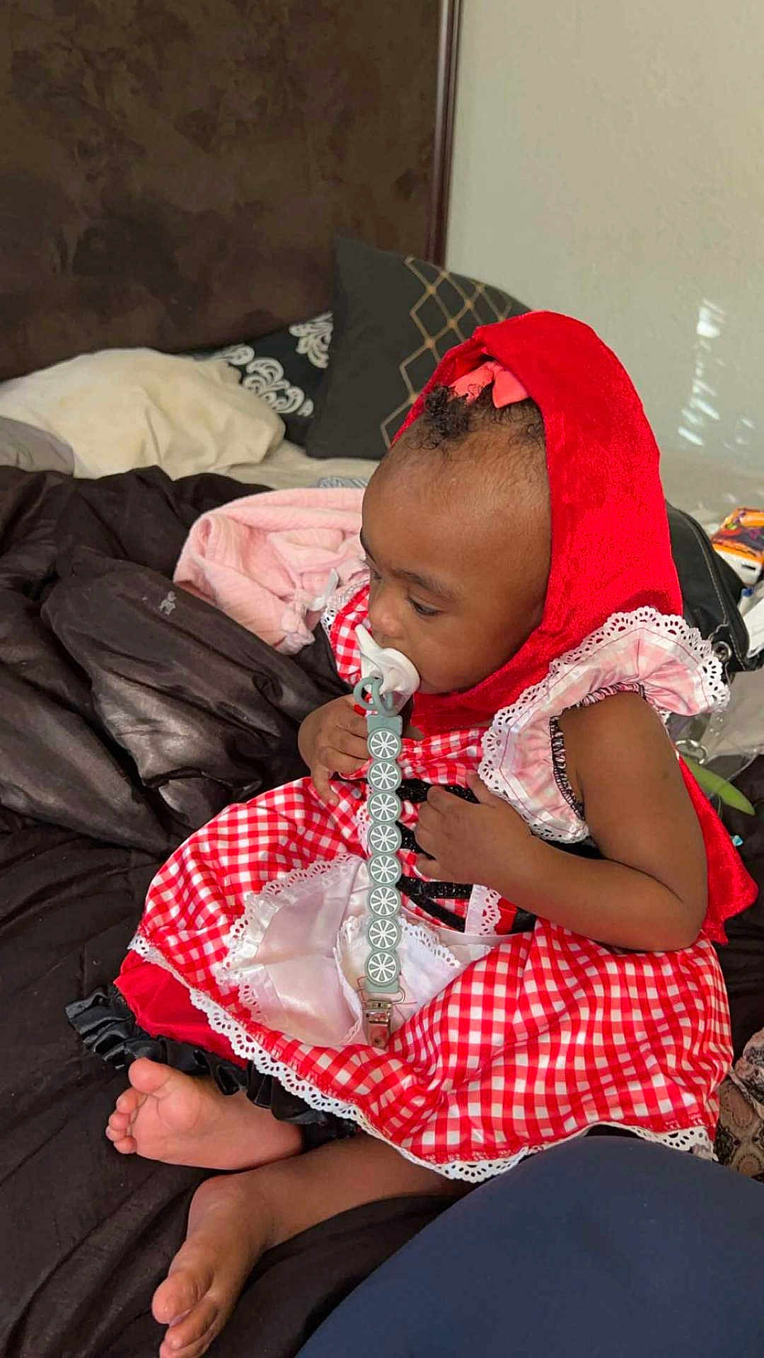 Delijah is registered to the contest to win money with this photo: toddler, child, baby, red_hood, pacifier, bed, blanket, pillow, dress, checkered_pattern, indoors, skin, barefoot, sitting, clothing, headwear, infant, cute, young_child, portrait