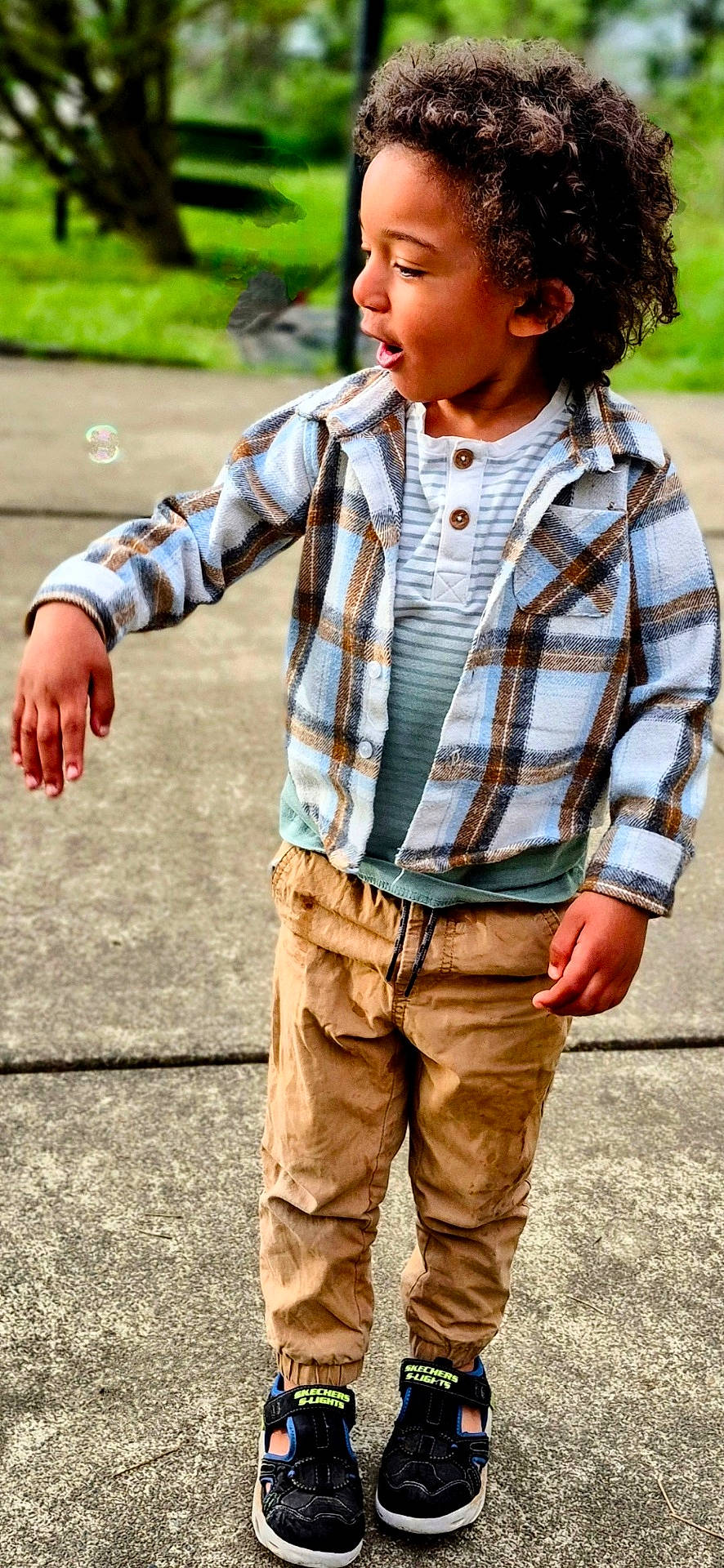 casual_clothing, child, concrete, curly_hair, daylight, expression, footwear, greenery, happy, kid, light_brown_pants, nature_background, outdoor, plaid_shirt, playful, side_profile, sneakers, standing, young