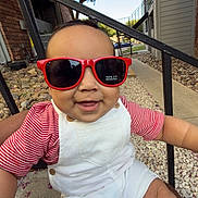Kyler is registered to the contest to win money with this photo: baby, child, sunglasses, red, white_overalls, striped_shirt, smiling, outdoor, residential_area, person, lap, sunlight, happy, face, head, arm, legs, rocks, tree, stairs