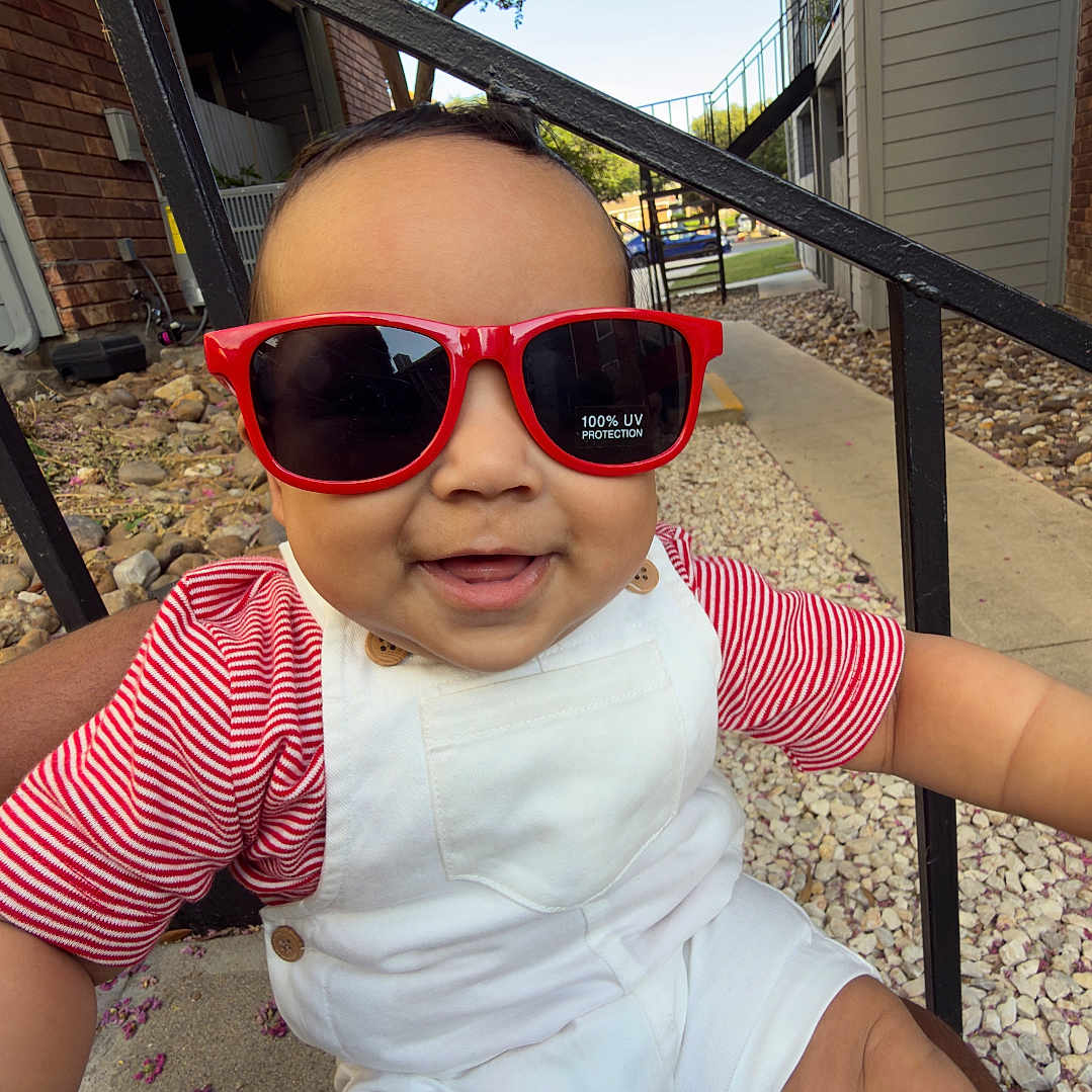 Kyler is registered to the contest to win money with this photo: baby, child, sunglasses, red, white_overalls, striped_shirt, smiling, outdoor, residential_area, person, lap, sunlight, happy, face, head, arm, legs, rocks, tree, stairs