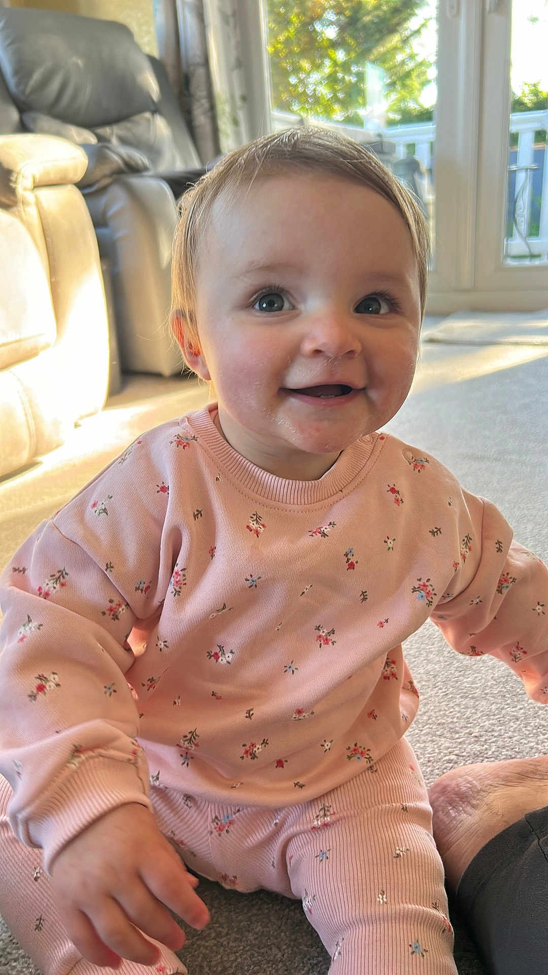 Ivy is registered to the contest to win money with this photo: baby, child, smiling, pink_clothing, floral_pattern, sitting, carpet, indoor, natural_light, sunlight, window, happy, cute, person, young_child, casual_wear, home_interior, cozy, portrait, candid