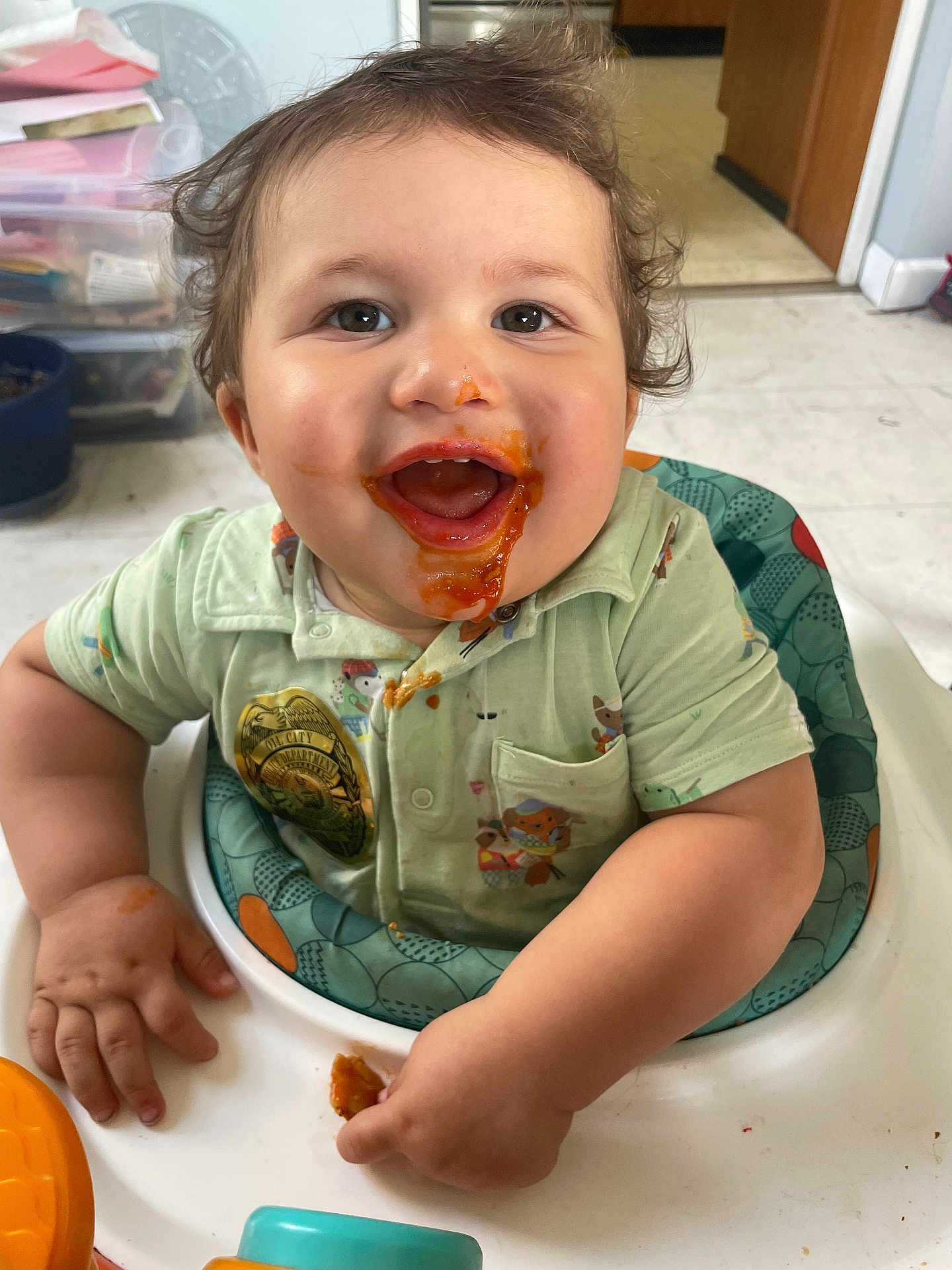 Amir joined the competition — help win amazing prizes! baby, child, messy_face, food, high_chair, smiling, happy, indoor, cute, clothing, green_shirt, hand, table, face, mouth_open, eyes, hair, kitchen, person, toddler