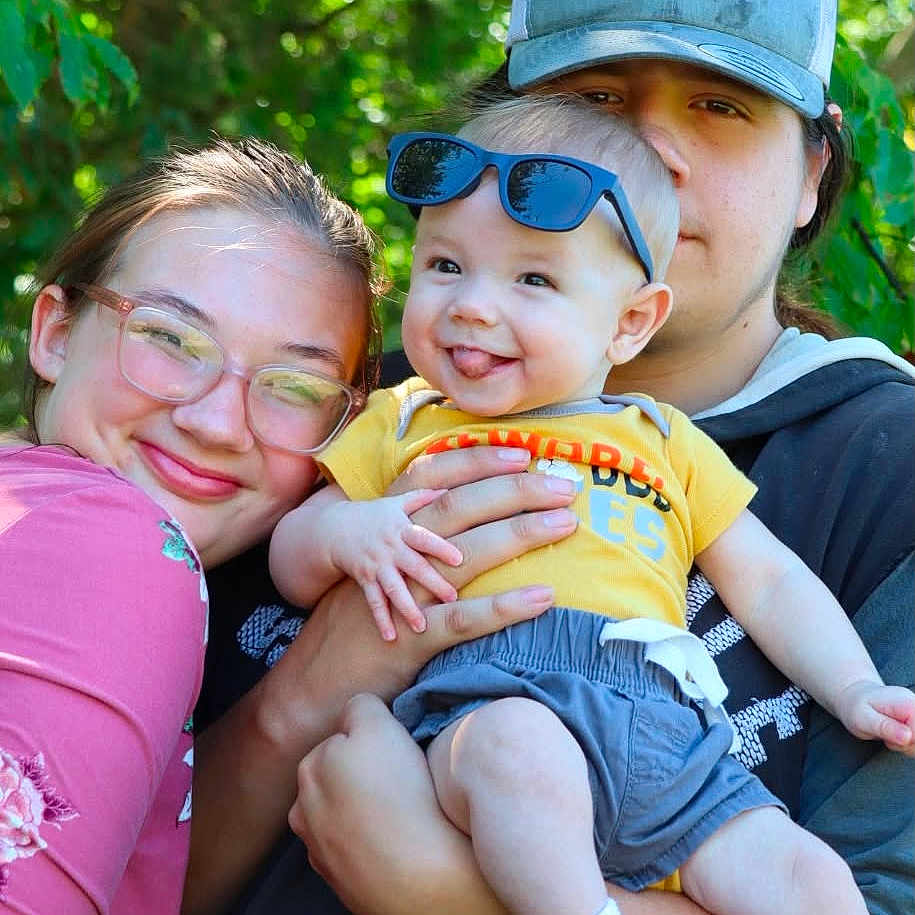 Bruce is registered to the contest to win money with this photo: baby, child, adult, family, outdoor, greenery, sunglasses, smile, happy, person, hat, glasses, clothing, nature, portrait, casual, holding, tongue_out, cute, toddler