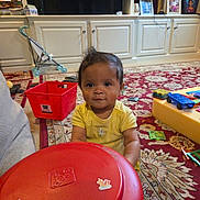 Kennedy is registered to the contest to win money with this photo: baby, infant, child, portrait, sitting, looking_at_camera, wide_eyes, smiling, yellow_shirt, playroom, living_room, toy_table, toy_truck, toy_stroller, storage_bin, red_table, patterned_rug, cabinet, indoors, family_home
