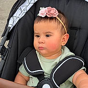 Abigail is registered to the contest to win money with this photo: baby, child, stroller, headband, flower, green_clothing, face, portrait, infant, pout, sitting, seatbelt, outdoor, curious, young_child, black_stroller, closeup, earrings, soft_lighting, serious_expression
