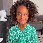 Leyla a rejoint le concours — aidez-le/la à gagner de superbes lots ! child, smile, curly_hair, green_dress, floral_pattern, indoors, living_room, couch, television, floor_tiles, portrait, happy, person, casual_clothing, young, cute, girl, standing, bright, natural_light