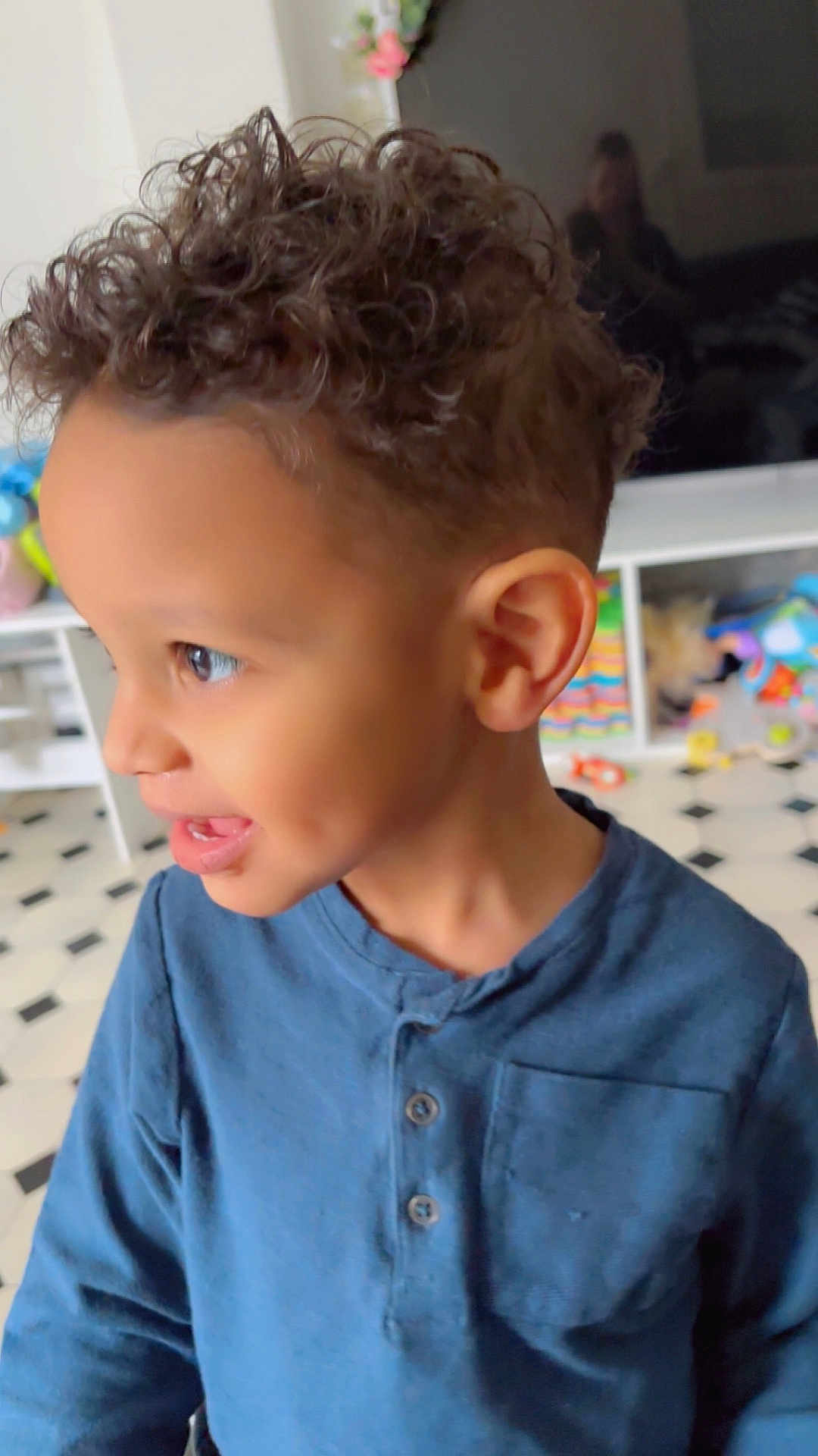 Noah a rejoint le concours — aidez-le/la à gagner de superbes lots ! child, curly_hair, dimples, blue_shirt, smiling, indoor, toys, playroom, happy, side_profile, young_child, person, floor_tiles, cabinet, television, reflection, colorful, home, casual_clothing, curly_haircut