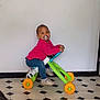 toddler, child, indoor, toy, tricycle, pacifier, pink_clothing, blue_pants, floor, tile_floor, patterned_floor, white_wall, shoes, play, riding, young_child, happy, person, baby, fun