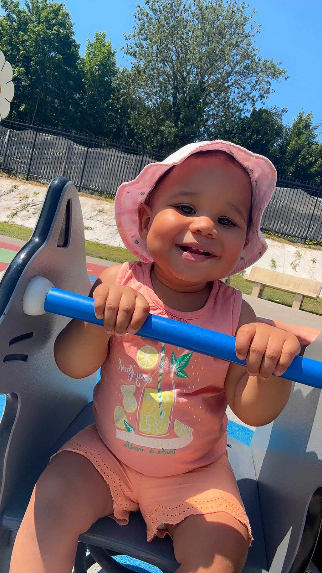 Zeyna a rejoint le concours — aidez-le/la à gagner de superbes lots ! toddler, child, smile, pink_hat, pink_clothing, playground, play_equipment, blue_bar, outdoor, sunny, trees, fence, bench, happy, holding, seat, summer, cute, baby, fun