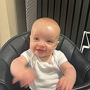 Finn is registered to the contest to win money with this photo: baby, child, high_chair, smile, happy, indoor, clothing, white_shirt, face, hands, waving, seat, black, furniture, background, dress, sparkly, human, infant, cute