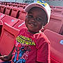 child, boy, smiling, cap, tshirt, spiderman, stadium, seats, red, sunlight, outdoor, happy, casual_clothing, portrait, person, daylight, young, cheerful, leisure, seating