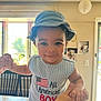 toddler, child, boy, smiling, hat, denim_hat, indoors, kitchen, table, holding_hands, person, window, sunlight, furniture, shirt, text_on_clothing, happy, portrait, casual, cute