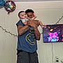 baby, man, holding, balloon, television, puppet, tattoo, smile, indoor, wall, clothing, shorts, tshirt, child, celebration, string_lights, power_strip, furniture, room, person