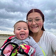 Elliott is registered to the contest to win money with this photo: baby, child, woman, glasses, beach, cloudy_sky, blanket, smiling, tongue_out, portrait, outdoor, sand, casual_clothing, person, family, headshot, happy, nature, fun, young_adult