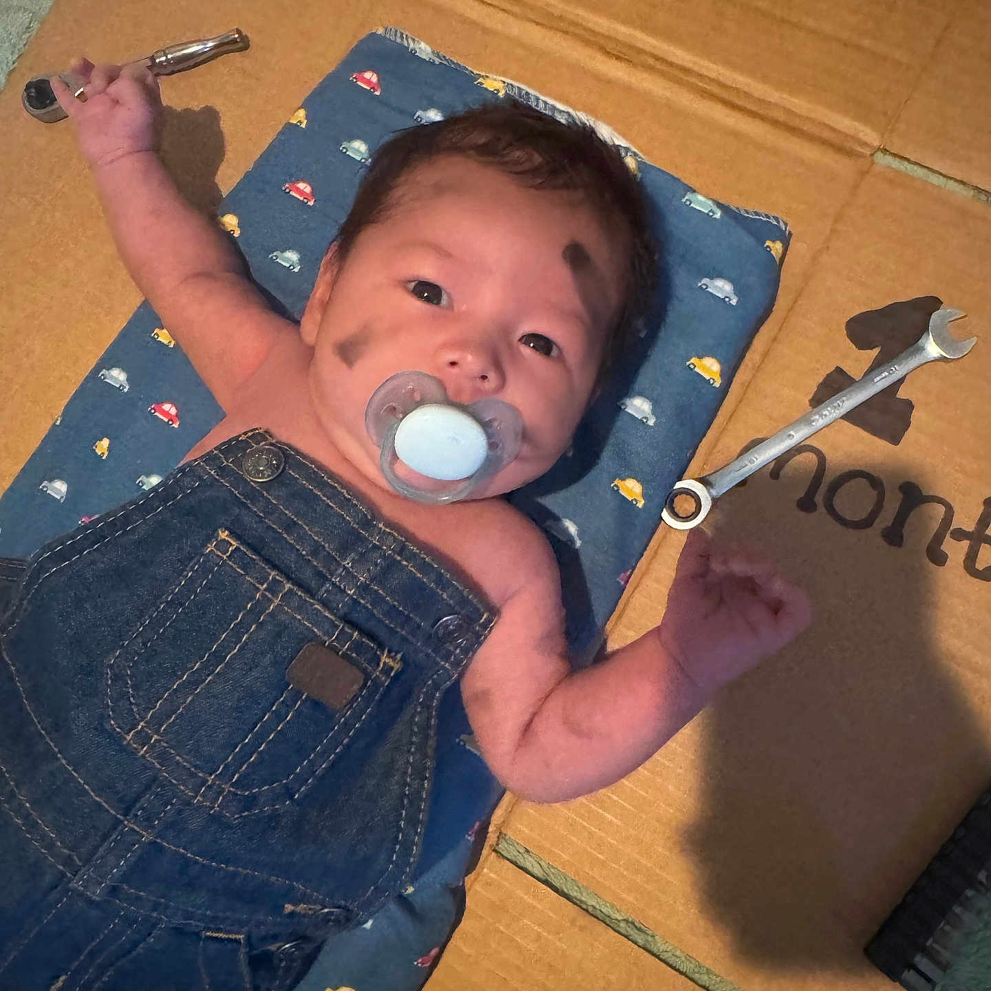 Liam is registered to the contest to win money with this photo: baby, cardboard, child, close_up, cute, denim_overalls, expression, face_paint, hands_up, indoors, infant, lying_down, mat, messy, pacifier, playful, portrait, tools, toy_cars_pattern, wrench