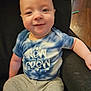 baby, infant, child, smiling, onesie, text_on_shirt, tie_dye, blue_shirt, sitting, arm, leg, sofa, couch, portrait, face, indoors, wood_floor, spoon, gray_pants, new_to_the_crew