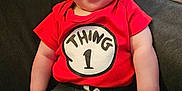 Nathan is registered to the contest to win money with this photo: arms, baby, child, chubby_cheeks, closeup, clothing, couch, face, happy, indoor, infant, legs, onesie, portrait, red_shirt, sitting, smiling, sofa, thing_1, wooden_floor