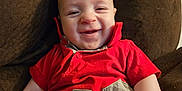 Nathan joined the competition — help win amazing prizes! baby, infant, child, smile, grin, red_shirt, couch, brown_fabric, face, eyes, cheeks, hand, arm, seated, plaid_pattern, number_patch, button, indoors, close_up, happy