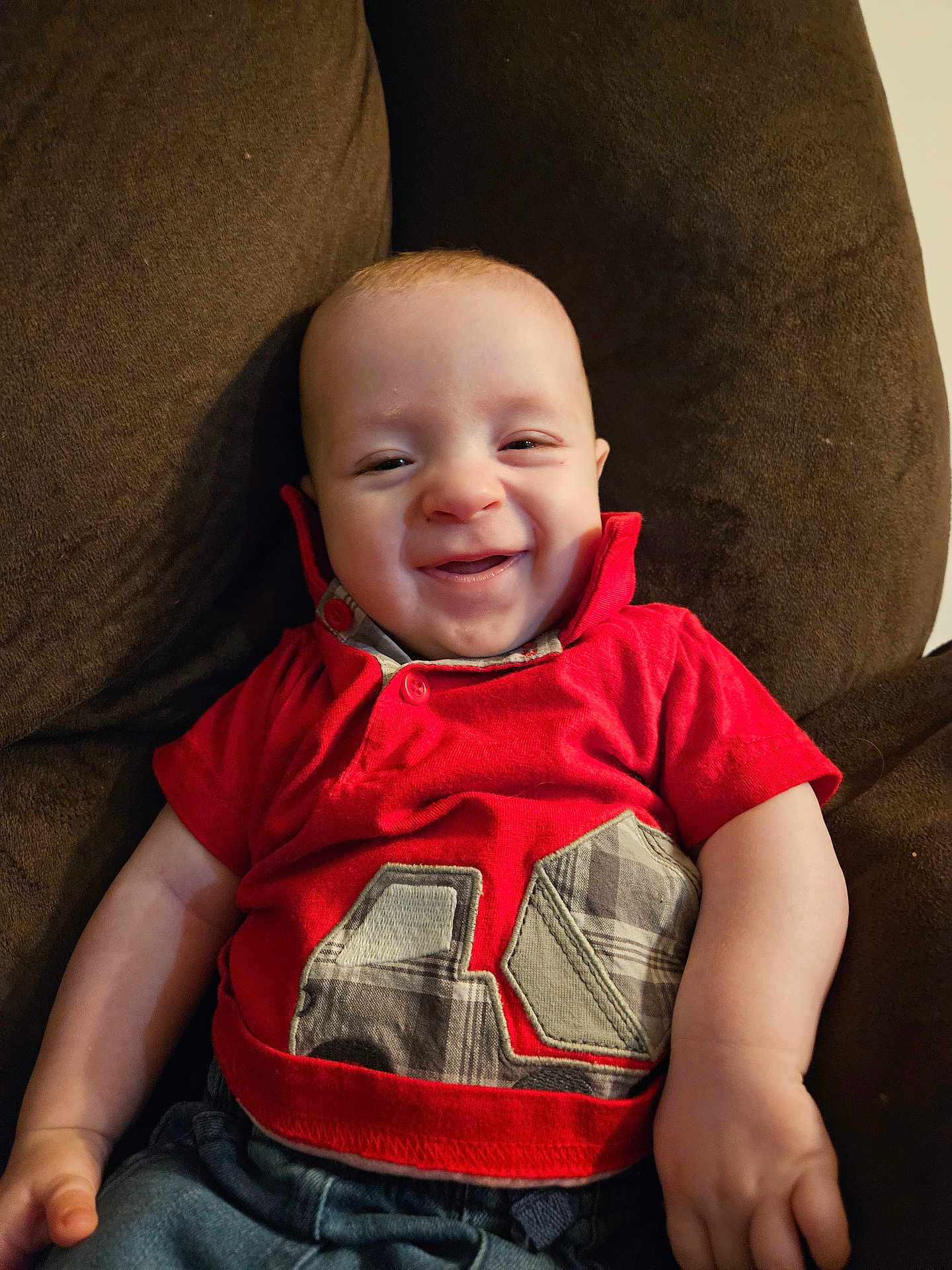 Nathan joined the competition — help win amazing prizes! baby, infant, child, smile, grin, red_shirt, couch, brown_fabric, face, eyes, cheeks, hand, arm, seated, plaid_pattern, number_patch, button, indoors, close_up, happy