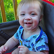 Nathan is registered to the contest to win money with this photo: baby, child, smiling, blue_eyes, short_hair, outdoor, grass, wooden_railings, shirt, blue_shirt, patterned_clothing, sitting, chair, red_strap, happy, portrait, cute, infant, sunlight, nature