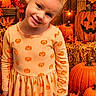 child, toddler, girl, smiling, portrait, autumn, halloween, pumpkins, jack_o_lantern, orange_dress, leaves, hay_bale, warm_lighting, indoors, fall_decorations, festive, head_tilt, blonde_hair, standing, photography