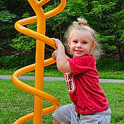 Paisley is registered to the contest to win money with this photo: child, playground, spiral_structure, yellow, red_shirt, gray_shorts, smiling, blonde_hair, hair_bun, outdoor, grass, trees, park, play, happy, person, young_child, casual_clothing, daylight, fun