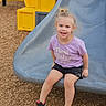 child, smiling, playground, slide, purple_shirt, shorts, sneakers, outdoor, wood_chips, yellow_play_equipment, happy, blond_hair, pony_tail, casual_clothing, sitting, daylight, fun, person, cute, youth