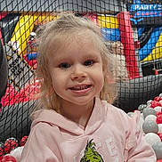 Paisley is registered to the contest to win money with this photo: child, smiling, hoodie, grinch, blonde_hair, indoor_play_area, ball_pit, net, red_balls, white_balls, gray_balls, playground, cute, happy, portrait, casual_clothing, young_child, fun, colorful, indoor