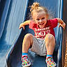 child, slide, playground, red_shirt, blonde_hair, smiling, happy, outdoor, colorful_shoes, blue_socks, shorts, sunlight, grip, fun, play, recreation, young, active, casual_clothing, daytime