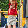 child, playground, slide, smile, red_shirt, gray_shorts, outdoor, greenery, trees, yellow_bars, play_equipment, happy, person, summer, fun, cute, topknot, casual_clothing, daylight, shoes
