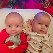 Isabella is registered to the contest to win money with this photo: baby, christmas_tree, cute, face, hands, holiday, indoor, infant, laying_down, onesie, ornament, patterned_clothing, pillow, portrait, red_sleeves, sibling, smiling, soft_lighting, twins, two_children