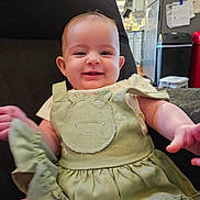 Isabella is registered to the contest to win money with this photo: baby, child, smiling, green_dress, face, arm, chair, indoor, kitchen, refrigerator, notes, furniture, happy, person, portrait, clothing, cute, infant, sitting, home