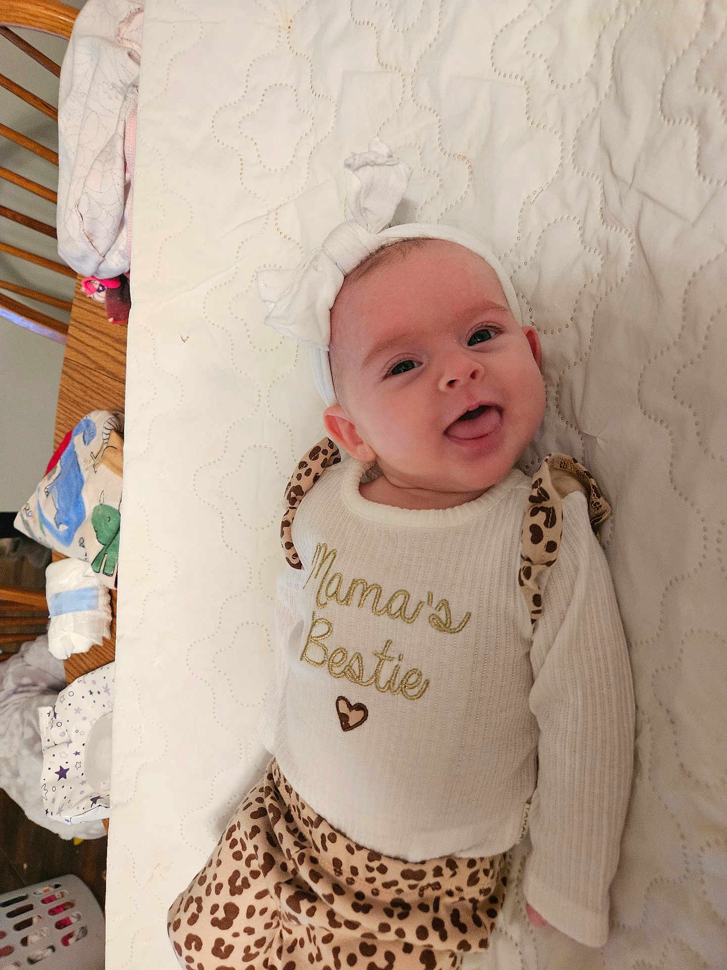 Isabella is registered to the contest to win money with this photo: baby, infant, smile, tongue_out, headband, bow, onesie, text_on_clothing, leopard_print, blanket, mattress, quilt, crib, wooden_furniture, diapers, nursery, clothing, portrait, cute, happy