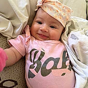 Valentina is registered to the contest to win money with this photo: baby, infant, smile, pink_clothing, hat, blanket, cushion, lying_down, cute, happy, child, face, portrait, soft_texture, indoors, cozy, headwear, small_person, rest, comfort