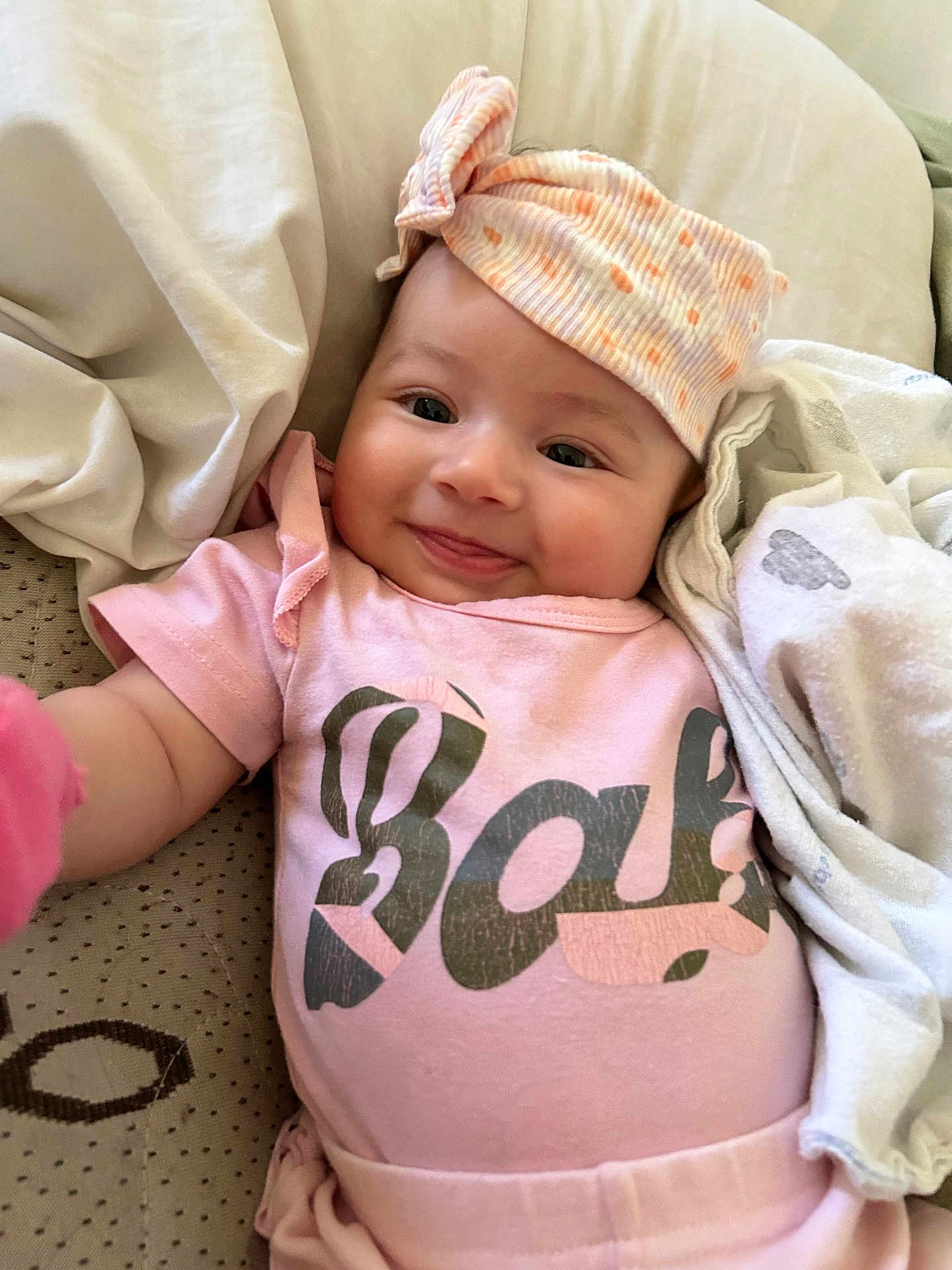 Valentina is registered to the contest to win money with this photo: baby, infant, smile, pink_clothing, hat, blanket, cushion, lying_down, cute, happy, child, face, portrait, soft_texture, indoors, cozy, headwear, small_person, rest, comfort