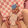 baby, smiling, blue_bow, headband, floral_outfit, chair, blanket, checkered_pattern, knit_throw, pink, beige, happy, portrait, infant, cute, sitting, indoors, child, person, cozy