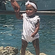 Honey is registered to the contest to win money with this photo: child, toddler, baby, outdoor, poolside, water, swimming_pool, white_outfit, dress, headband, bow, sandals, standing, holding_hand, adult_hand, portrait, sunlight, dock, rocks, happy